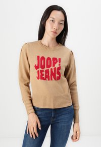 Beige gebreide trui met geribbelde manchetten en zoom, met een opvallend rood "JOOP! JEANS" logo op de voorkant. Draagt met donkerblauwe jeans.