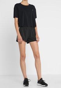 Henkilö, jolla on päällä löysä musta lyhythihainen t-paita, mustat urheilushortsit ja mustat juoksukengät, seisoo valkoista taustaa vasten.