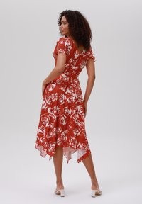 Robe fleurie rouge avec des manches courtes, dos en V et ourlet asymétrique. Fabriquée en matériau léger avec une superposition transparente et un motif de roses blanches.