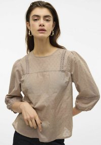 Blusa marrone chiaro con maniche a palloncino, tessuto testurizzato con piccoli punti e dettagli in pizzo lungo le spalle e i polsini.