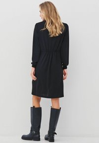 Černé šaty s dlouhými rukávy, nabíraným pasem, délka ke kolenům, doplněné vysokými černými botami. Hladká textura a jednoduchý design.