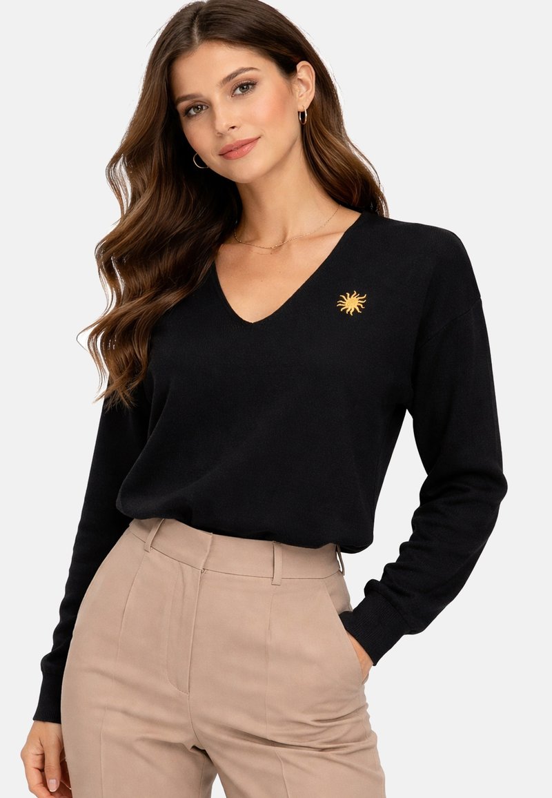 Femme aux longs cheveux bruns ondulés portant un pull noir à col en V avec un petit emblème de soleil jaune et un pantalon beige à taille haute.