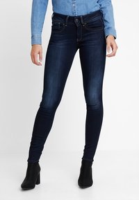 Jeans ajustados de color azul oscuro, confeccionados en denim con una textura suave, que presentan costuras decorativas, bolsillos delanteros y un cierre de botón.