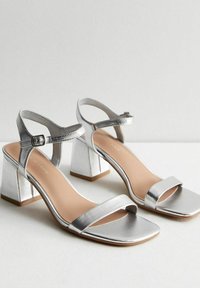 Silver metallic klackskor med fyrkantig tå, justerbar ankelrem och blockklack. Slät textur med en minimalistisk design.