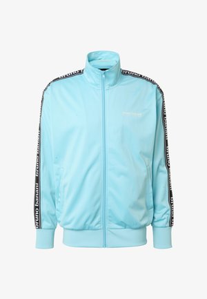 Hele sinine lukustatav trennijakk kõrge kraega, mustade küljeribadega, millel on "bruno banani" tekst, ja kahe ees oleva taskuga.