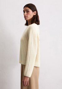 Cremfarbener Strickpullover mit vertikalen Mustern, Rundhalsausschnitt und gerippten Bündchen. Kombiniert mit beigen, hochgeschnittenen Hosen.