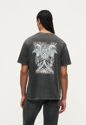 Mand med fletninger iført falmet sort T-shirt med grafiske englevinger og teksten "Replay Black" og "Electro Thunder" på ryggen.