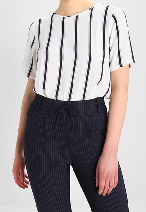 Femme portant une blouse blanche à manches courtes avec des rayures noires verticales et un pantalon bleu marine à fines rayures avec une taille à cordon.