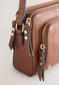 Sac à main en cuir brun texturé avec fermetures éclair et garnitures dorées. Comprend des détails tressés, plusieurs compartiments et des accents noirs contrastants.