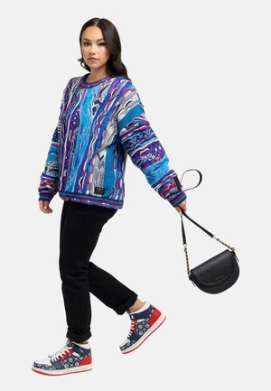 Frau trägt einen bunten blau gemusterten Pullover, schwarze Hose und gemusterte Sneakers, hält eine schwarze Handtasche am Trageriemen.