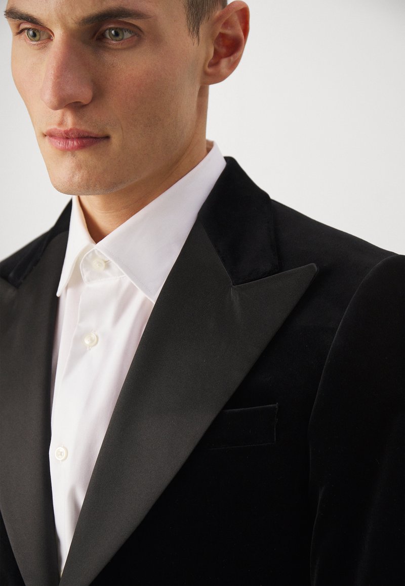 Czarna aksamitna marynarka smokingowa z satynowymi klapami w szpic, zestawiona z białą koszulą wizytową z klasycznym kołnierzykiem i zapięciem na guziki.