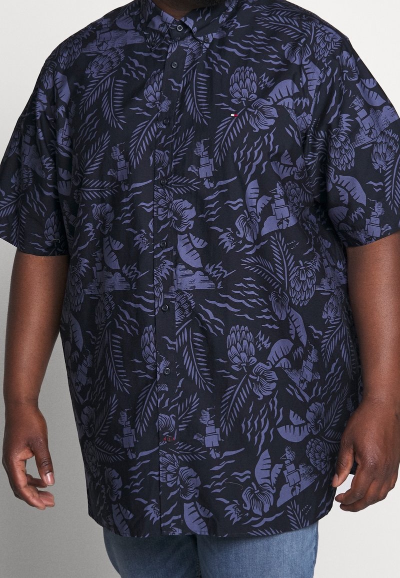 Chemise à manches courtes avec boutons, arborant des motifs floraux tropicaux et des feuilles de couleur bleu foncé et violet, portée par une personne.