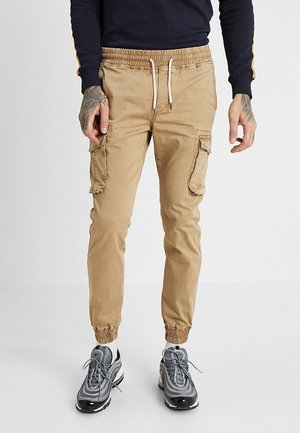 Mann trägt beige Cargo-Jogginghosen mit Kordelzugbund, elastischen Bündchen, navyblauen Pullover und graue Sportschuhe, steht auf weißem Boden.