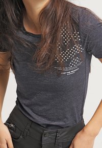 Camiseta de manga corta gris oscuro hecha de material suave, con un patrón de puntos blancos y texto en la parte superior izquierda. Diseño ajustado con cuello redondo.