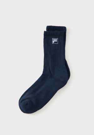 Une chaussette unique bleu marine de style crew avec le haut côtelé et un petit logo blanc et bleu marine près du bord supérieur sur un fond clair uni.