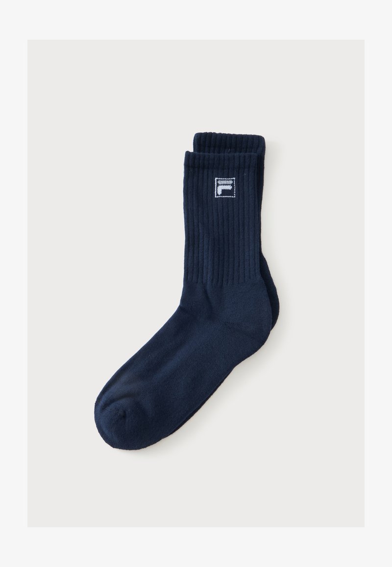Une chaussette unique bleu marine de style crew avec le haut côtelé et un petit logo blanc et bleu marine près du bord supérieur sur un fond clair uni.