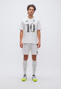 Νεαρός άνδρας στέκεται φορώντας λευκή φανέλα ποδοσφαίρου με τον αριθμό 10, το λογότυπο της Γιουβέντους, τα λογότυπα της Adidas και νέον κίτρινα παπούτσια ποδοσφαίρου σε απλό φόντο.