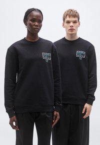 Sudaderas negras con un parche de logo en turquesa, con puños y bajos acanalados, usadas por dos modelos sobre un fondo neutro.