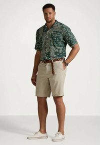 Man i grönmönstrad kortärmad skjorta, beige shorts med flätat bälte, vita sneakers och klocka, står med handen i fickan.
