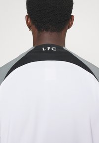 Camisa deportiva blanca con acentos en negro y gris, presentando el logo "LFC" en la zona del cuello. Textura suave y ajuste atlético.