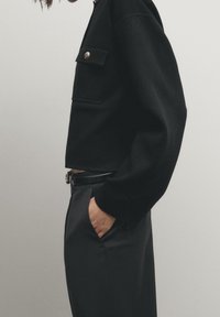 Svart kortärmad tröja med bröstficka och knappdetalj, tillsammans med hög midja svarta byxor. Släta texturer och rena linjer.