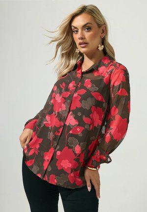 Blonde vrouw draagt een doorschijnende zwarte blouse met knopen en grote rode en grijze bloemenpatronen, gecombineerd met zwarte broek en gouden oorbellen.
