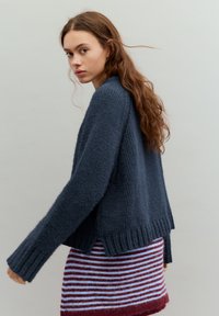 Tengerészkék kötött pulóver széles, bordázott szegéllyel és eresztett vállvarrással, csíkos bordó és világoskék szoknya fölött viselve.