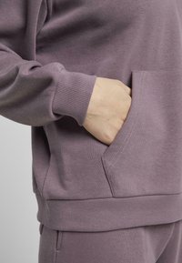Lawendowa bluza z ściągaczami, z kangurzą kieszenią. Materiał wydaje się miękki i lekko teksturowany.