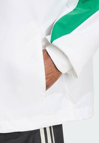 Giacca bianca con accenti verdi, dotata di tasca con zip e polsini elasticizzati. Materiale liscio e leggero. Design casual per abbigliamento attivo.