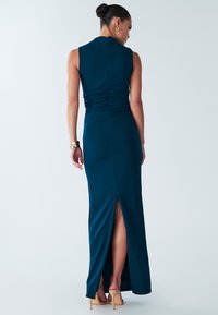 Groene, aansluitende maxi-jurk met een hoge halslijn, gerimpelde achterkant en een hoge split aan de dij. Gemaakt van gladde stof. Gecombineerd met neutrale hakken.