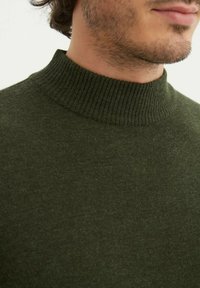 Pull en tricot vert avec un col montant côtelé, présentant un tissu doux et texturé ainsi qu'une coupe ajustée. Aucun motif ni accent visible.