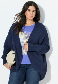 Marinblå oversize stickad cardigan med texturerad yta, öppen framtill och ribbade manschetter, lager-på-lager över en blå grafisk T-shirt och denimjeans.