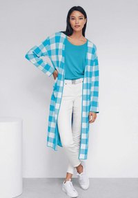 Lange open voorkardigan in een blauw-wit geruit patroon, gecombineerd met een effen turquoise top en witte jeans, aangevuld met witte sneakers.