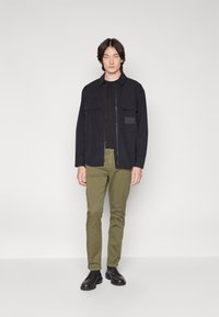 Giacca nera con zip e due tasche frontali sopra una maglietta nera, abbinata a pantaloni verdi slim-fit e stivali neri stringati. Tessuto dalla consistenza liscia.