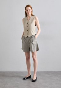 Beige mouwloos vest met knoopsluiting, gecombineerd met grijze geplooide shorts. Zwarte ballerina's accentueren de outfit. Minimalistisch ontwerp, gladde stof.