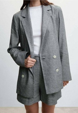 Kvinde iført en grå dobbeltradet blazer og matchende shorts med en hvid ribbet top under, stående mod en ensfarvet baggrund.