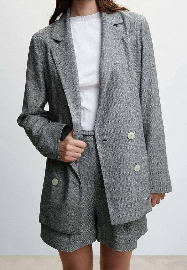 Femme portant un blazer gris à double boutonnage et un short assorti avec un haut blanc côtelé en dessous, debout devant un fond uni.