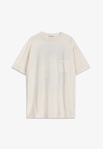 T-shirt à manches courtes de couleur crème avec un col rond et une petite poche poitrine sur le côté gauche, posé à plat sur un fond blanc.