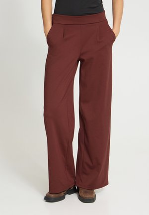 Personne portant un pantalon marron à jambes larges, les mains dans les poches et des bottes marron visibles en bas sur un fond uni.