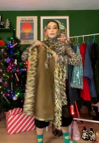 Mujer sosteniendo un abrigo marrón con forro de estampado de leopardo en una habitación con un árbol de Navidad, regalo envuelto y un perchero con ropa de colores.