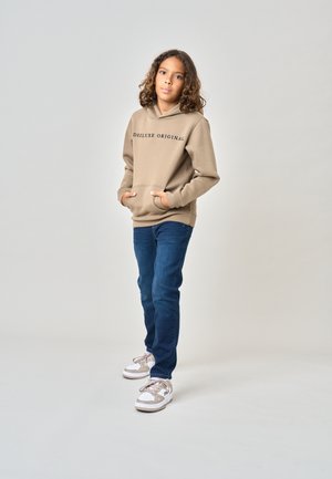 Enfant aux cheveux bouclés portant un sweat à capuche beige avec texte, jean bleu et baskets blanches, debout devant un fond clair uni.