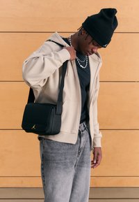 Svart skinn skulderveske med teksturert overflate, gullfarget logo, justerbar stropp. Bær over en beige hettegenser og grå jeans.