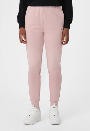 Persona che indossa pantaloni da jogging rosa chiaro e scarpe da ginnastica bianche, in piedi davanti a uno sfondo chiaro uniforme.