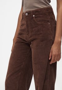 Gros plan d'une personne portant un pantalon en velours côtelé marron avec un bouton en argent, et un haut sans manches beige sur fond blanc.