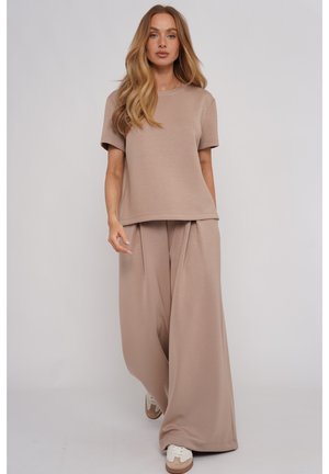 Kobieta o długich blond włosach, ubrana w luźny beżowy krótki top i szerokie spodnie, idzie do przodu w białych trampkach na jednolitym tle.