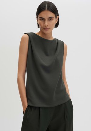 Donna con capelli corti e scuri che indossa una canotta grigio scuro senza maniche e pantaloni scuri abbinati, con le mani nelle tasche davanti a uno sfondo semplice.