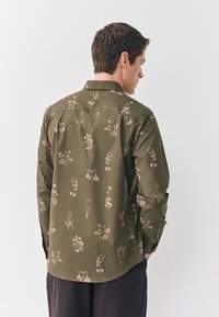 Oliivinvihreä paita, jossa vaaleanpunainen kukkakuvio. Paidassa on nappikaulus ja pitkät hihat, joissa napilliset mansetit, ja sileä pinta.