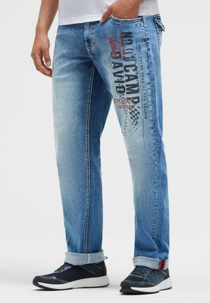 Mann trägt hellblaue Jeans mit grafischem Text am rechten Oberschenkel und hochgekrempelten Säumen, kombiniert mit schwarz-weißen Sneakers.