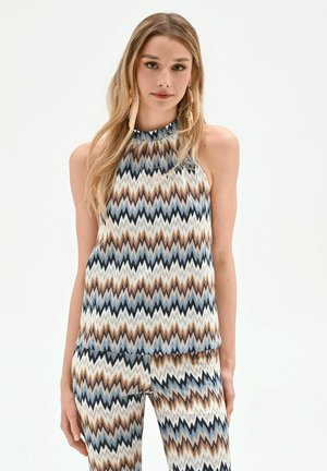 Femme portant un haut sans manches à col montant et un pantalon assorti avec un motif en zigzag dans des tons bleus, marron, noir et blanc, debout sur fond blanc.