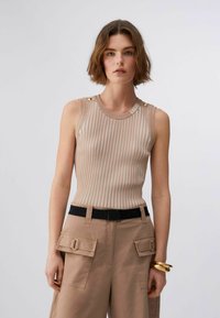 Jeune femme portant un haut sans manches beige et blanc à rayures avec des boutons dorés, un pantalon cargo beige, une ceinture noire et des bracelets dorés.